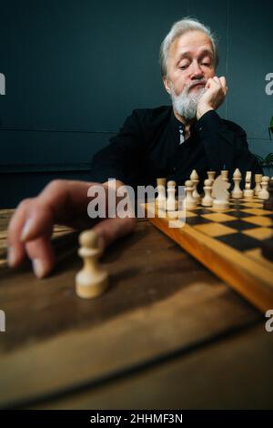 Vertikale Low-Angle-Aufnahme von nachdenklichen grauhaarigen älteren Mann denken Spielstrategie auf Holztisch mit Schachbrett sitzen, selektive Fokus. Stockfoto