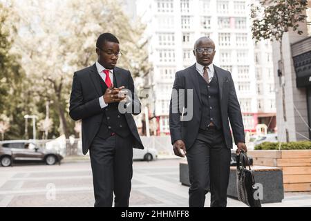 Zwei dunkelhäutige afroamerikanische Geschäftsleute in Anzügen und Brillen gehen in der Stadt draußen spazieren und diskutieren über Geschäfte Stockfoto