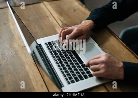 Nahaufnahme aus einem hohen Winkel des nicht erkennbaren älteren Mannes, der auf der Laptop-Tastatur am Tisch im Wohnzimmer sitzt. Stockfoto