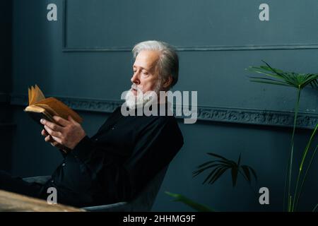 Fokussierter älterer grauhaariger älterer Mann, der zu Hause ein Papierbuch liest, sitzt am Tisch, entspannt sich und ruht sich mit Literatur aus. Stockfoto