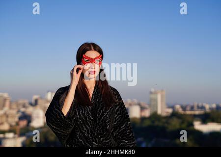 Porträt von niedlichen Brünette Superhelden Mädchen in schwarzem Kleid und roten Gesichtsmaske auf blauen Himmel und Stadtstadt Hintergrund. Frauenmacht, Frauenrechte, Protest- oder Aktivismus-Konzept. Hochwertige Bilder Stockfoto