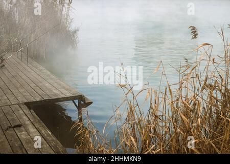 Ein Pier am Ufer des Sees mit Angelruten und trockenem Schilf im Herbst nebligen Wetter. Stockfoto