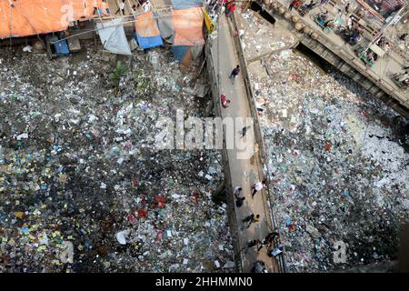 JANUAR 25,2022,DHAKA,BANGLADESH- die Menschen gehen an einer Brücke über ein verschmutztes Gebiet und Kinder gehen über ein verschmutztes Gebiet, während sie Plastikpaten sammeln Stockfoto