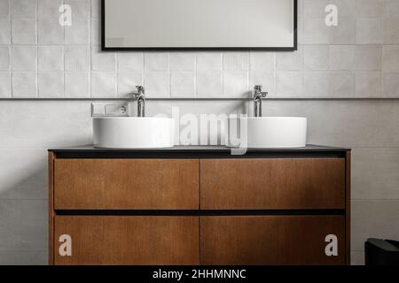 Badezimmer mit modernem Waschtisch mit zwei runden Keramikwaschbecken, Chromarmaturen und Spiegel mit schwarzem Rahmen, weißen Geschichten und Holzschrank. Moderne Ba Stockfoto