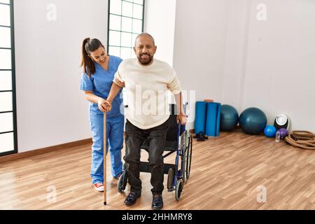 Älterer Mann, der auf dem Rollstuhl sitzt, wird von einer Altenpflegerin unterstützt, behinderter reifer Mann, der Hilfe beim Aufstehen bekommt Stockfoto