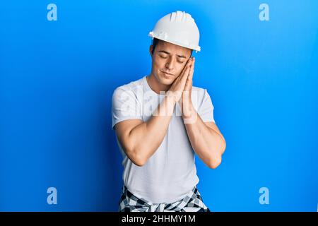 Hübscher junger Mann mit Baueruniform und Hardhut, müde schlafend, träumend und mit Händen zusammen posiert, während er mit geschlossenen Augen lächelt. Stockfoto