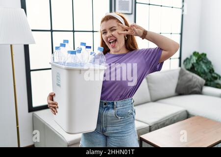 Junge Rotschopf Frau hält Recycling-Papierkorb mit Plastikflaschen tun Frieden Symbol mit Fingern über Gesicht, lächelnd fröhlich zeigt Sieg Stockfoto