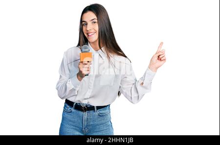 Junge Brünette Teenager halten Reporter Mikrofon lächelnd glücklich zeigt mit Hand und Finger zur Seite Stockfoto