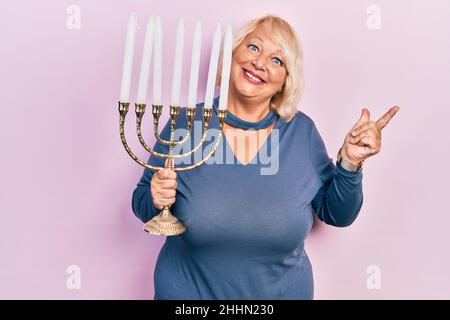 Blonde Frau mittleren Alters mit der Menorah hanukkah jüdische Kerze lächelnd glücklich zeigt mit Hand und Finger zur Seite Stockfoto