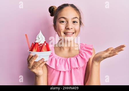 Schöne Brünette kleines Mädchen essen Erdbeer-Eis feiert Leistung mit glücklichen Lächeln und Gewinner Ausdruck mit erhobener Hand Stockfoto