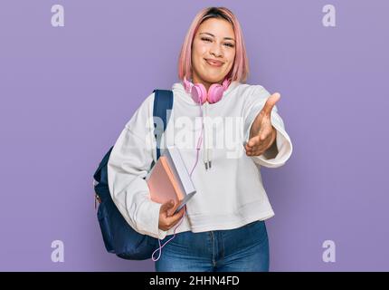 Hispanische Frau mit rosa Haaren, die einen Studentenrucksack und Kopfhörer trägt, lächelt fröhlich und bietet mit der Handfläche Unterstützung und Akzeptanz. Stockfoto