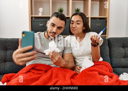 Junges lateinamerikanisches krankes Paar, das zu Hause eine medizinische Fernsultation mit dem Smartphone hat. Stockfoto