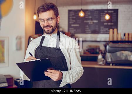 Fröhlicher männlicher Arbeiter, der im Café auf die Zwischenablage schrieb Stockfoto