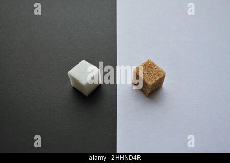 Konfrontation: Zwei Zuckerstücke - raffiniertes Weiß auf schwarzem Hintergrund und Rohrzucker auf weißem Hintergrund einander gegenüber. Stockfoto