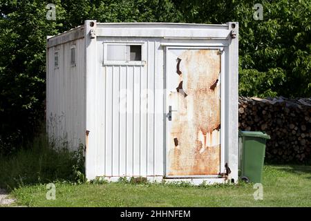 Weißer alter verlassene Industriecontainer, der in Lagerraum umgewandelt wurde, mit kleinen Fenstern und verrosteten, gebrochenen Türen vorne links im Vorort Stockfoto