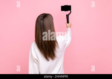 Rückansicht des weiblichen Livestreams, mit Standicam und Telefon, rückwärts vor der Kamera posiert, trägt weißen Pullover im lässigen Stil. Innenaufnahme des Studios isoliert auf rosa Hintergrund. Stockfoto