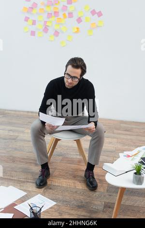 Trendiger Mann, der Dokumente in der Nähe von Papieren auf dem Boden und dem Couchtisch ansieht Stockfoto