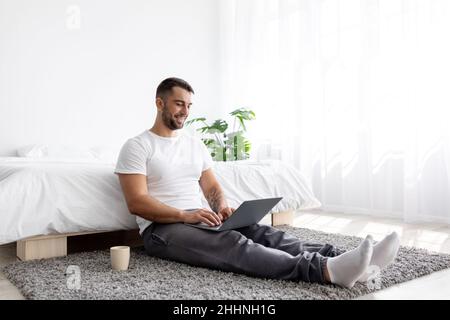 Lächelnder junger kaukasischer, hübscher, muskulöser Mann, der auf dem Boden sitzt und im Schlafzimmer auf dem Laptop tippt Stockfoto