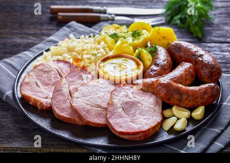 Gebratene Bratwurst, geräucherter Schweinehals, gekochte Kartoffeln, Sauerkraut und Senf auf schwarzem Teller, deutsche Küche, horizontale Ansicht von oben Stockfoto