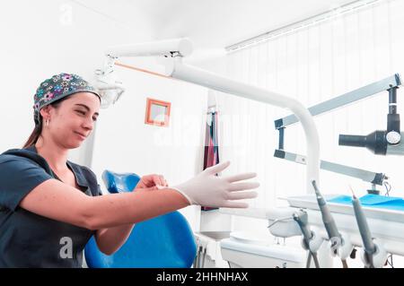 Bild eines hübschen Mädchens mit weißer Haut und professioneller Zahnärztin mit grauer Uniform und farbenfroher Mütze, die in ihrem Büro Latex-Handschuhe anlegt, um zu st Stockfoto