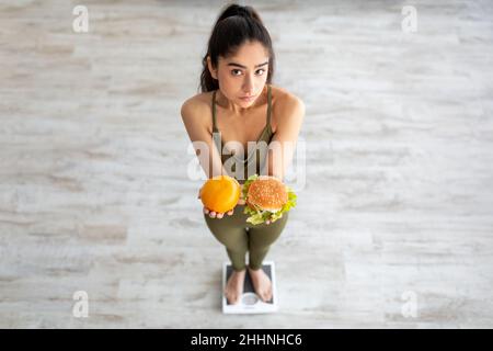 Oberhalb der Ansicht einer jungen Inderin, die Zitrusfrüchte und Hamburger hält, auf einer Waage steht und die Wahl getroffen hat Stockfoto