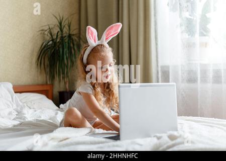 Happy Little lustige Mädchen verwendet Laptop für Videoanruf. Kaukasisches lockiges blondes Kind in Hasenohren studiert ferngesteuert Video über Laptop abspielen oder ansehen Stockfoto