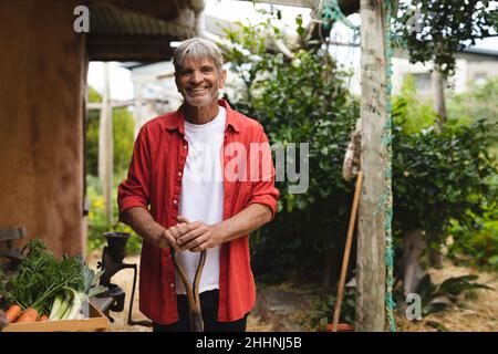 Portrait eines glücklichen kaukasischen reifen Mannes mit Schaufel, der von geerntetem Gemüse im Hinterhof steht. Lifestyle und Landwirtschaft. Stockfoto