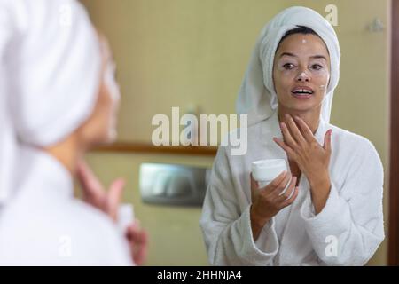 Biracial junge Frau trägt Bademantel Anwendung Feuchtigkeitscreme vor dem Spiegel im Badezimmer. Unverändert, Routine, Lifestyle und Vitiligo Konzept. Stockfoto
