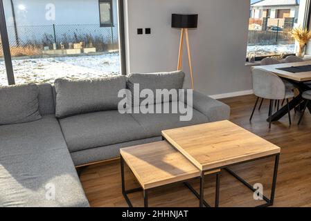 Ein modernes und einfaches Wohnzimmer im Haus mit einem grauen Sofa, sichtbaren großen Terrassenfenstern und zwei Tischen, die sich untereinander befinden. Stockfoto