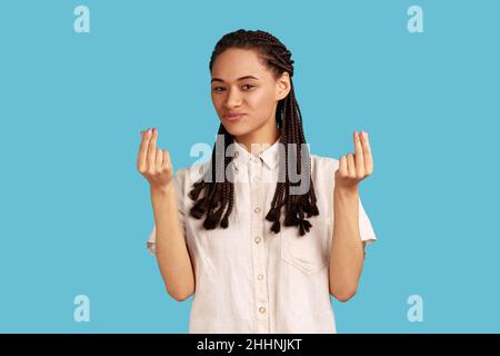 Porträt einer schönen Frau mit schwarzen Dreadlocks macht Geld Geste, reibt Finger, Blick auf die Kamera mit froher Ausdruck, trägt weißes Hemd. Innenaufnahme des Studios isoliert auf blauem Hintergrund. Stockfoto