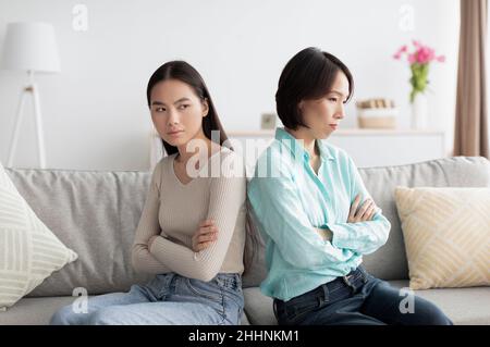 Beleidigte die junge asiatische Frau und ihre reife Mutter, die sich nach einem Streit auf der Couch zurücklehnen und sich gegenseitig ignorieren Stockfoto