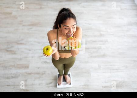 Cheat Meal Konzept. Oben die tausendjährige indische Dame, die auf einer Waage steht, Apfel hält und Hamburger isst Stockfoto