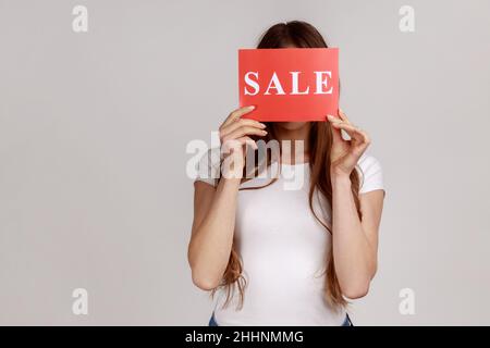 Porträt einer anonymen, unbekannten jungen Frau, die ihr Gesicht hinter einer Karte versteckt, mit Aufschriften zum Verkauf, Einkaufen, Rabatten, weißem T-Shirt. Innenaufnahme des Studios isoliert auf grauem Hintergrund. Stockfoto