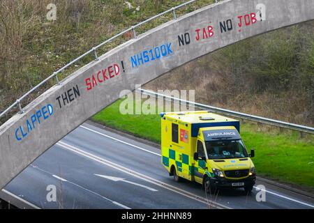 Weymouth, Dorset, Großbritannien. 25th. Januar 2022. Ein Krankenwagen des NHS fährt am A354 in Weymouth in Dorset unter einer Brücke durch, auf der eine Graffiti-Nachricht geschrieben wurde, in der stand: „klatscht dann geplündert! NHS100K Keine Jabs Keine Jobs! Die Botschaft ist erschienen, da die Frist für die obligatorische Covid-Impfung der NHS-Mitarbeiter durch die Regierungen mit der Frist für den ersten Jab am 3rd. Februar und für die vollständige Impfung bis zum 1st. April 2022 anrückt. Bildnachweis: Graham Hunt/Alamy Live News Stockfoto