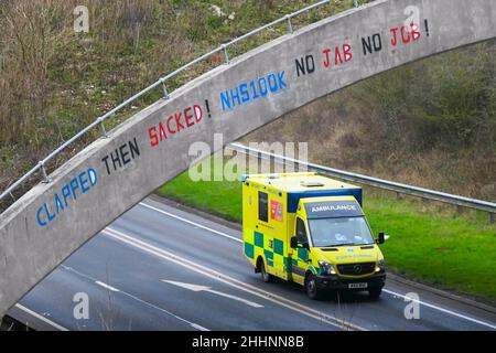 Weymouth, Dorset, Großbritannien. 25th. Januar 2022. Ein Krankenwagen des NHS fährt am A354 in Weymouth in Dorset unter einer Brücke durch, auf der eine Graffiti-Nachricht geschrieben wurde, in der stand: „klatscht dann geplündert! NHS100K Keine Jabs Keine Jobs! Die Botschaft ist erschienen, da die Frist für die obligatorische Covid-Impfung der NHS-Mitarbeiter durch die Regierungen mit der Frist für den ersten Jab am 3rd. Februar und für die vollständige Impfung bis zum 1st. April 2022 anrückt. Bildnachweis: Graham Hunt/Alamy Live News Stockfoto