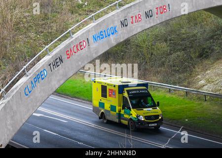 Weymouth, Dorset, Großbritannien. 25th. Januar 2022. Ein Krankenwagen des NHS fährt am A354 in Weymouth in Dorset unter einer Brücke durch, auf der eine Graffiti-Nachricht geschrieben wurde, in der stand: „klatscht dann geplündert! NHS100K Keine Jabs Keine Jobs! Die Botschaft ist erschienen, da die Frist für die obligatorische Covid-Impfung der NHS-Mitarbeiter durch die Regierungen mit der Frist für den ersten Jab am 3rd. Februar und für die vollständige Impfung bis zum 1st. April 2022 anrückt. Bildnachweis: Graham Hunt/Alamy Live News Stockfoto