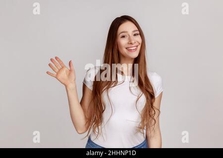 Hallo. Porträt einer glücklichen Frau, die mit erhobener Hand zur Kamera steht und die Gäste mit einem freundlichen Lächeln begrüßt und ein weißes T-Shirt trägt. Innenaufnahme des Studios isoliert auf grauem Hintergrund. Stockfoto