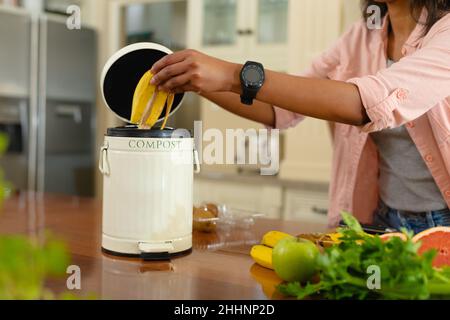 Mittelteil einer jungen afroamerikanischen Frau, die zu Hause Bananenschalen in Kompostbehälter legt. Menschen und Komposttechnologie-Konzept, unverändert. Stockfoto