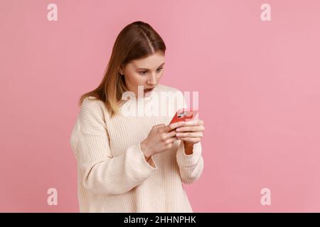 Porträt einer schönen, erstaunt blonden Bloggerin, die Smartphone benutzt, auf den Bildschirm des Geräts schaut, schockierende Inhalte sieht, trägt einen weißen Pullover. Innenaufnahme des Studios isoliert auf rosa Hintergrund. Stockfoto