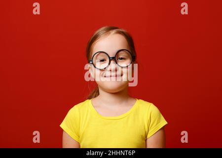 Lustige kleine Mädchen mit Brille. Schulmädchen Kind in einem gelben T-Shirt auf einem roten isolierten Hintergrund. Stockfoto