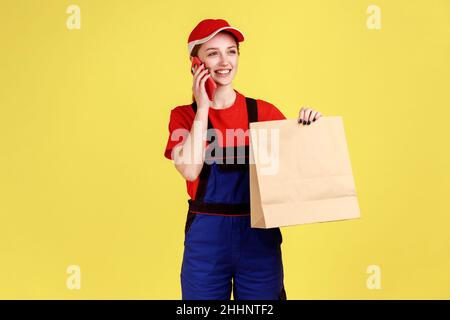 Porträt einer lächelnden Kurierin, die mit einem Papierpaket in den Händen steht und mit dem Kunden telefoniert, Overalls und roter Mütze trägt. Innenaufnahme des Studios isoliert auf gelbem Hintergrund. Stockfoto