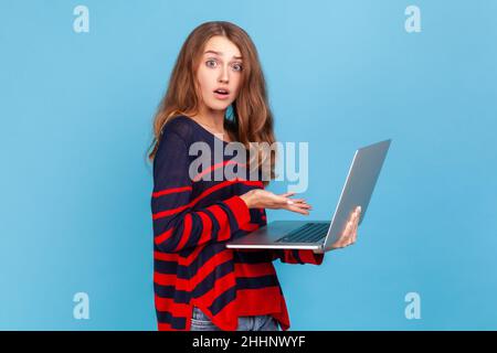 Staunende junge Frau, die einen gestreiften Pullover im lässigen Stil trägt, einen Laptop in den Händen hält, schockiert über die Anforderungen an die Arbeit und die Frist. Innenaufnahme des Studios isoliert auf blauem Hintergrund. Stockfoto