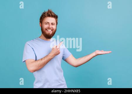 Produktwerbung. Porträt eines entzückten bärtigen Mannes, der auf den Kopierraum auf der Handfläche zeigt und einen leeren Platz für kommerzielle Texte oder Waren zeigt. Innenaufnahme des Studios isoliert auf blauem Hintergrund. Stockfoto