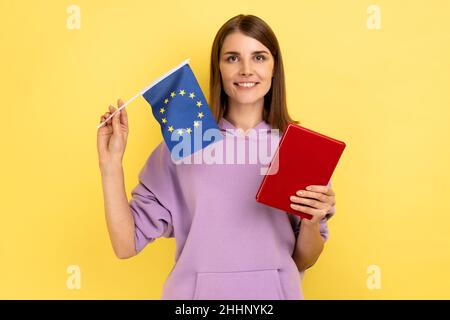 Porträt einer lächelnden, schönen Studentin mit Büchern und der Flagge der Europäischen Union, Bildung im Ausland, mit purpurfarbenem Hoodie. Innenaufnahme des Studios isoliert auf gelbem Hintergrund. Stockfoto