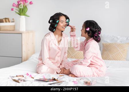 Fröhliches chinesisches Teenager-Mädchen mit Lockenwicklern im Haar wendet Kosmetik auf das Gesicht einer tausendjährigen Frau in rosa Pyjama an Stockfoto