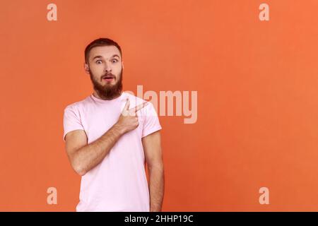 Porträt eines staunenden bärtigen Mannes, der mit einem überraschten Gesicht auf die Kamera schaute und auf den Wandkopierraum zeigte, der ein rosa T-Shirt trug. Innenaufnahme des Studios isoliert auf orangefarbenem Hintergrund. Stockfoto