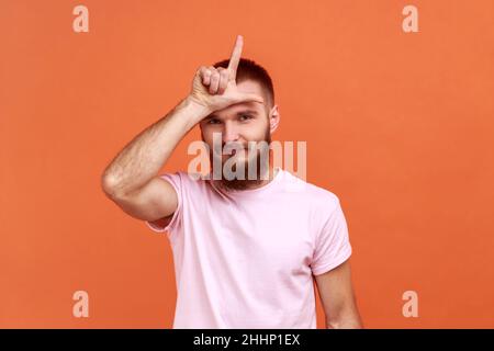 Porträt eines bärtigen Mannes, der die Hand an die Stirn hält, mit einer loseren Geste mit den Fingern, mit finanziellen und geschäftlichen Problemen und einem rosa T-Shirt. Innenaufnahme des Studios isoliert auf orangefarbenem Hintergrund. Stockfoto