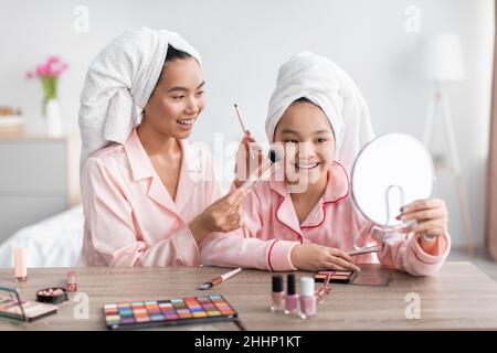 Fröhliche asiatische junge Dame und Teenager-Mädchen in rosa Pyjamas Anwendung Make-up und Blick auf Spiegel im Schlafzimmer Stockfoto