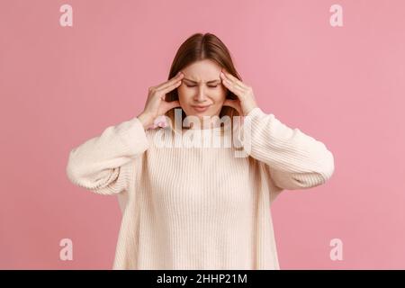 Porträt einer ungesunden, kranken, blonden Frau, die Schläfen massiert und mit Händen schreckliche Kopfschmerzen, Migränegefahr und weißen Pullover trägt. Innenaufnahme des Studios isoliert auf rosa Hintergrund. Stockfoto