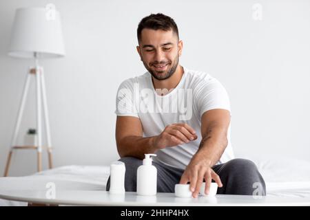 Lächelnder junger kaukasischer muskulöser, hübscher Mann am Tisch mit Gläsern verschiedener feuchtigkeitsspendender Anti-Aging-Kosmetik Stockfoto